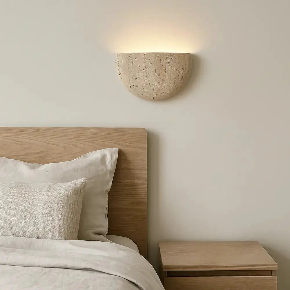 Applique murale demi-lune en travertin naturel diffusant une lumière douce au-dessus d’un lit dans une chambre minimaliste.